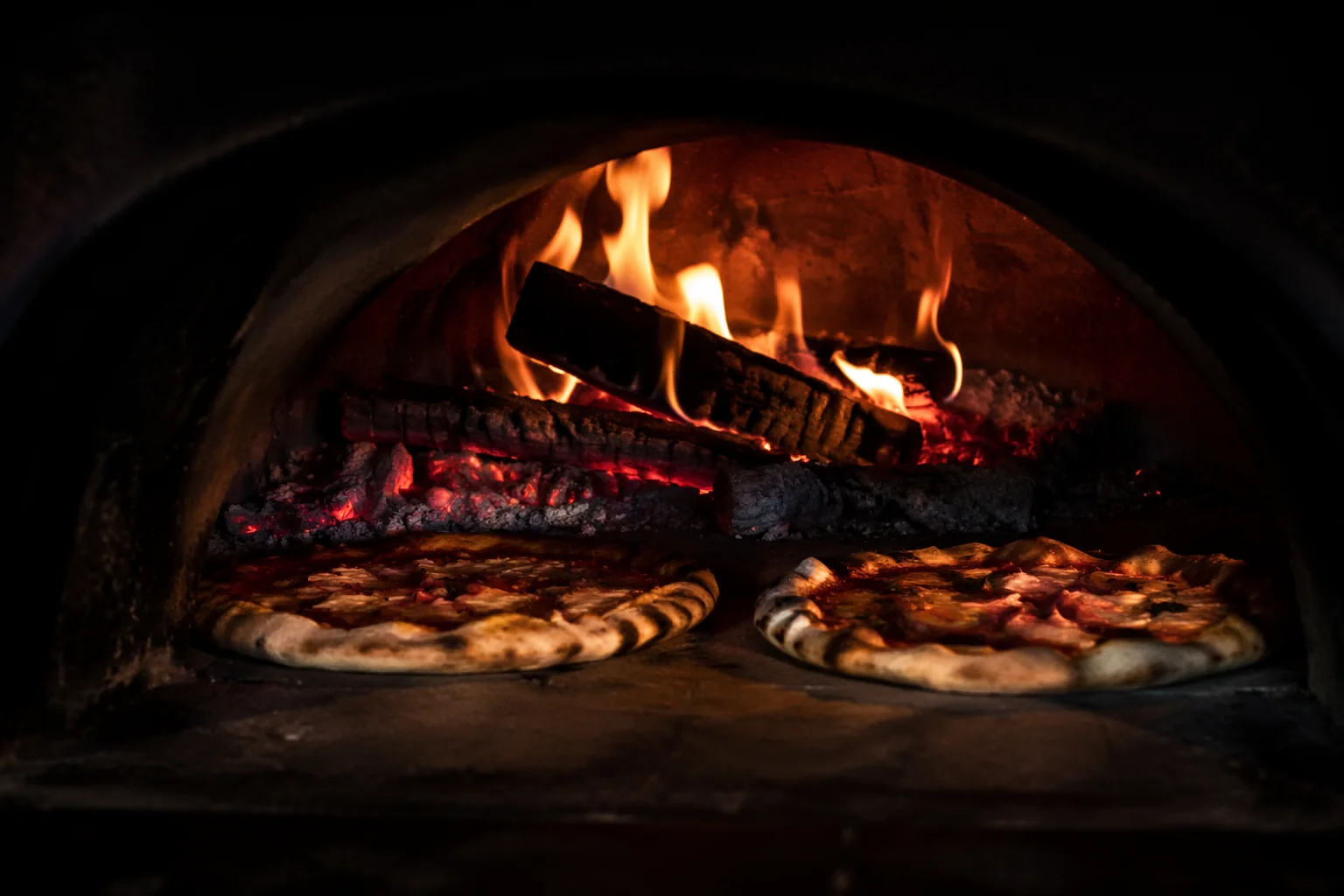 Flames rolling over split red oak logs inside the brick pizza oven, two pizzas baking at the oven mouth