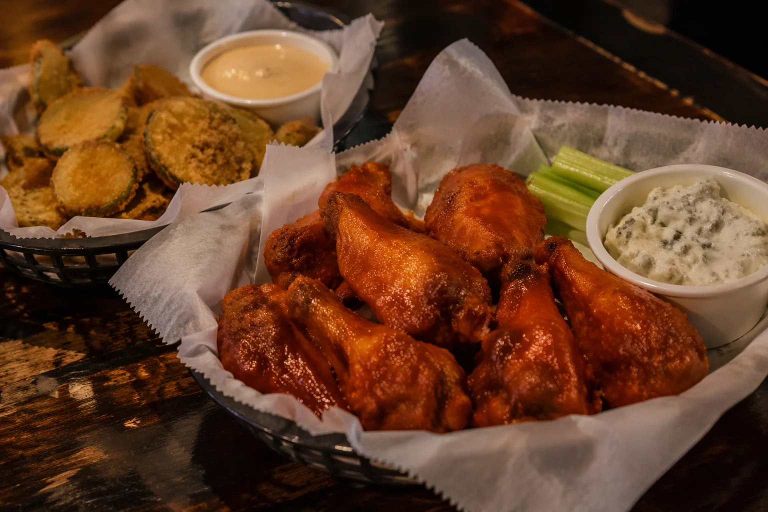 Basket of ten buffalo wings in glossy sauce with celery and blue cheese dip, fried pickle slices with chipotle ranch behind, on a dark wood table